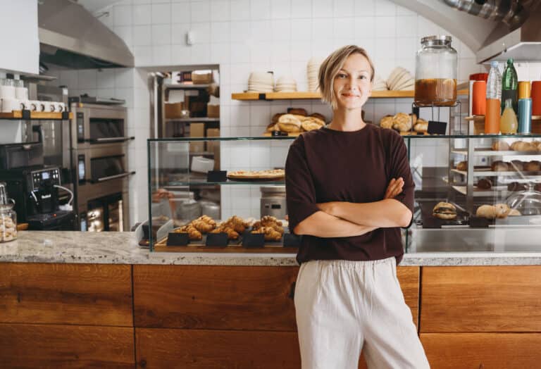 Portrait of smiling young woman entrepreneur standing at the counter of her bakery and coffee shop. Local small business owner indoors. Female in cafe near showcase with fresh croissants and bread.