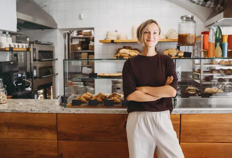 Portrait of smiling young woman entrepreneur standing at the counter of her bakery and coffee shop. Local small business owner indoors. Female in cafe near showcase with fresh croissants and bread.