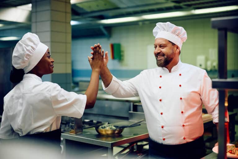 Happy chef giving high five to his African American cook assistant while working in the kitchen.